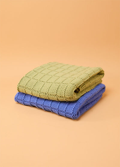Two folded Window Pane Blankets in green and blue, stacked against a beige background, each featuring a textured square panel design.
