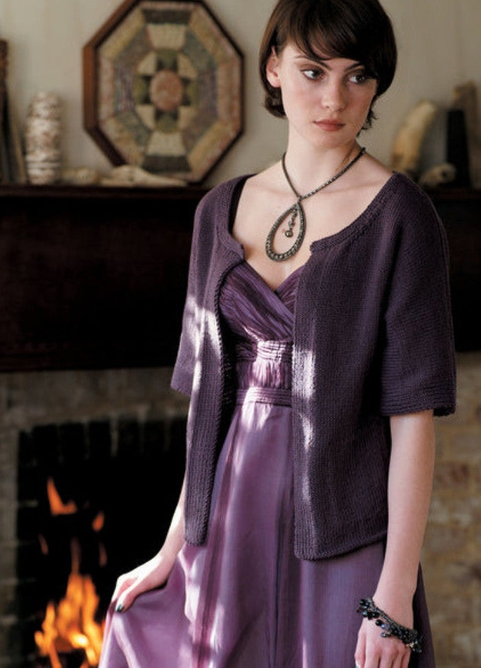 Young woman in a purple dress and cardigan stands by a fireplace, looking pensive. The room has warm tones, with a decorative wall piece above the mantle.