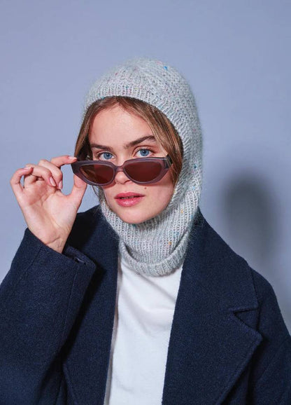 A person wearing a knitted grey hood and a navy coat adjusts stylish brown sunglasses, exuding a cool, confident vibe against a soft blue background.