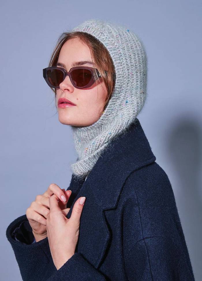 Woman in a blue coat with a knitted hood and sunglasses against a light blue background, conveying a stylish and confident tone.