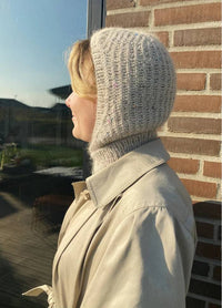 A person in a beige coat and knitted hood stands in profile, lit by warm sunlight. They're beside a brick wall and glass window, conveying a serene, cozy mood.