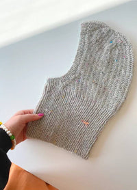 A hand with pink nail polish holds a knitted grey balaclava with coloured speckles, lying on a white surface. The mood is cozy and handmade.
