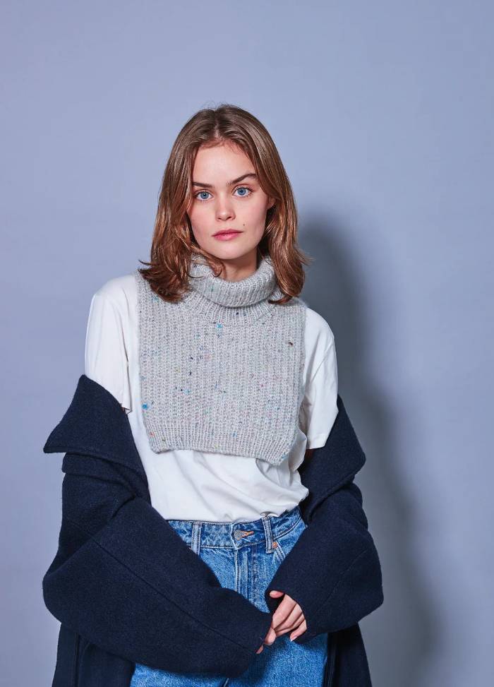 Young woman poses in a casual outfit against a light blue background. She wears a grey turtleneck neck warmer over a white top, layered with a dark coat, and jeans.