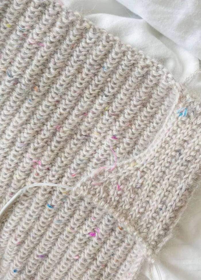 Close-up of a hand-knitted beige fabric with colourful specks on a soft white background. The texture is ribbed and intricate, conveying warmth.