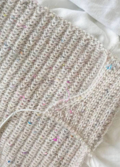 Close-up of a hand-knitted beige fabric with colourful specks on a soft white background. The texture is ribbed and intricate, conveying warmth.