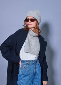 A woman in sunglasses wears a grey knit hat and neck warmer, navy coat, and jeans. She stands confidently against a light blue background, exuding a cool, casual vibe.