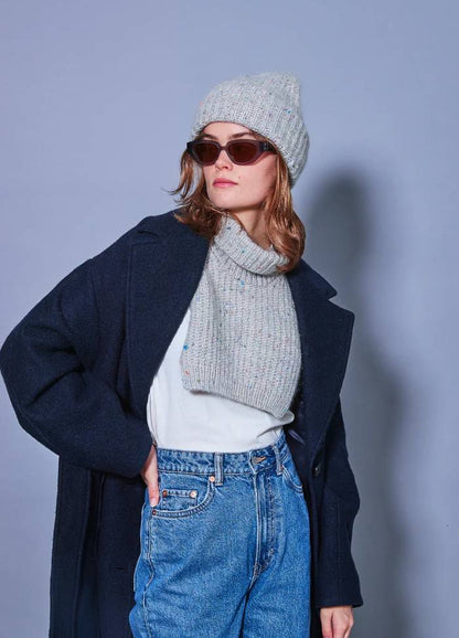 A woman in sunglasses wears a grey knit hat and neck warmer, navy coat, and jeans. She stands confidently against a light blue background, exuding a cool, casual vibe.
