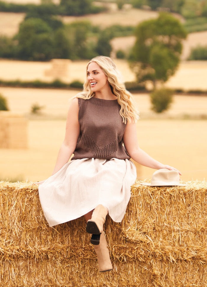 Woman in a brown sleeveless top and white skirt sits joyfully on a hay bale, holding a hat. Rural landscape with trees and fields.