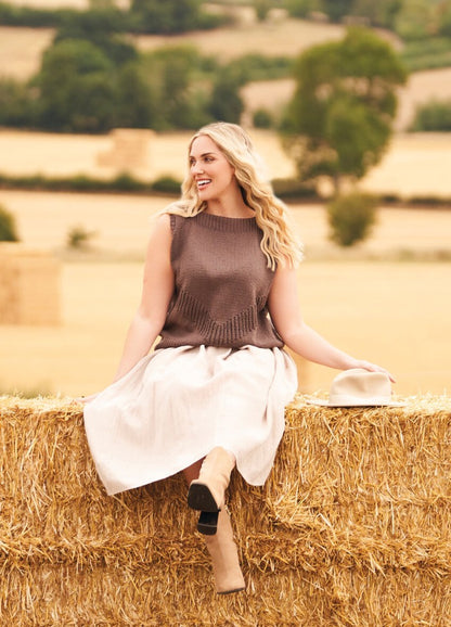 Woman in a brown sleeveless top and white skirt sits joyfully on a hay bale, holding a hat. Rural landscape with trees and fields.