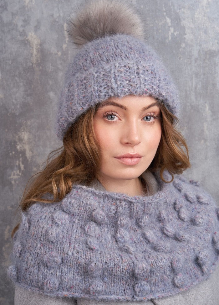 Woman wearing a knitted gray hat with a fur pom-pom and matching shawl. Neutral expression, soft lighting, textured gray background. Cozy and stylish.