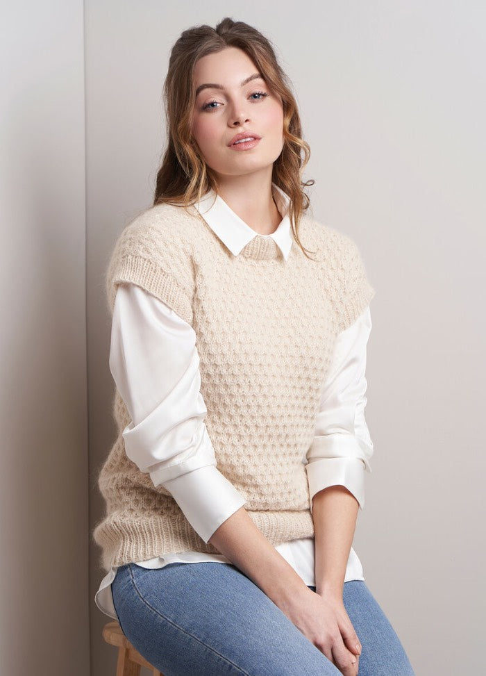Young woman with wavy brown hair, wearing a beige knit vest over a white blouse, and jeans, sits against a neutral backdrop, exuding a relaxed, confident vibe.